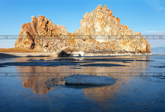 Spring morning on Baikal Lake. View of the famous Shamanka rock - a natural landmark of Olkhon Island in the rays of the rising sun. The bay is covered with a thin crust of blue ice. Scenic spring landscape. Стоковое фото, фотограф Виктория Катьянова / Фотобанк Лори
