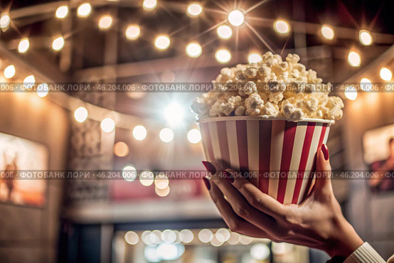 Striped red and white box with salted popcorn in his hand against the background of cinema lights. A popular snack in cinemas. Стоковое AI-изображение, AI-художник Photojuli / Фотобанк Лори