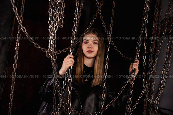 Stylish classy teen girl model 12-13 year posing in dark industrial room with chains, looking at camera. Cute chic teenage girl actress in leather jacket. Fashionable image concept. Copy ad text space. Стоковое фото, фотограф Aleksandr Golubev / Фотобанк Лори