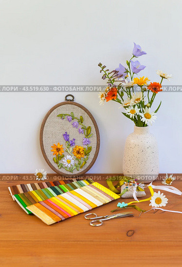 Summer still life with handmade floral embroidery with satin ribbons of flowers in hoop. Bouquet of daisies, marigolds and blue bells on wooden table. Crafts and embroidery as hobby. DIY concept. Стоковое фото, фотограф Виктория Катьянова / Фотобанк Лори