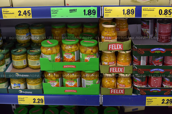 Supermarket shelves with canned vegetables. In the foreground are glass jars of pickled pumpkin from the Freshona and Felix brands. The products are neatly laid out in cardboard boxes with the company logos. In the background, other canned goods are visible: cucumbers, salads, and cans of Campolargo beans. Above the shelf are bright yellow and green price tags indicating the cost of the goods. The scene illustrates the atmosphere of a grocery store, the variety of products, and the design of the shelves with goods. (2015 год). Редакционное фото, фотограф Анна Кривицкая / Фотобанк Лори