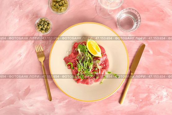 Тарелка с вкусным карпаччо из телятины, стакан воды, каперсы и консервированный халапеньо на цветном фоне. Стоковое фото, фотограф Pixel_shot / Фотобанк Лори