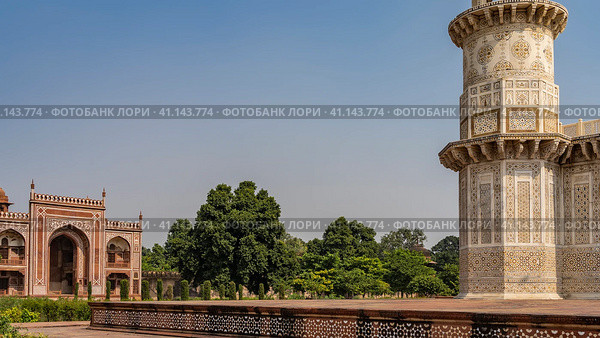 The ancient complex tomb of Itmad-Ud-Daulah. Marble corner minaret with ornaments, inlays of precious stones against the blue sky. In the distance is a red sandstone building with arches. India. Agra (2022 год). Стоковое фото, фотограф Вера Ивановна Кубарева / Фотобанк Лори