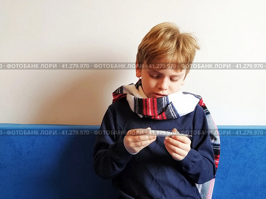 The boy got sick, his throat hurts and his body temperature rises. The childs neck is wrapped in a warm scarf. The kid looks dejectedly at the thermometer and is surprised. Стоковое фото, фотограф Юлия Петровская / Фотобанк Лори