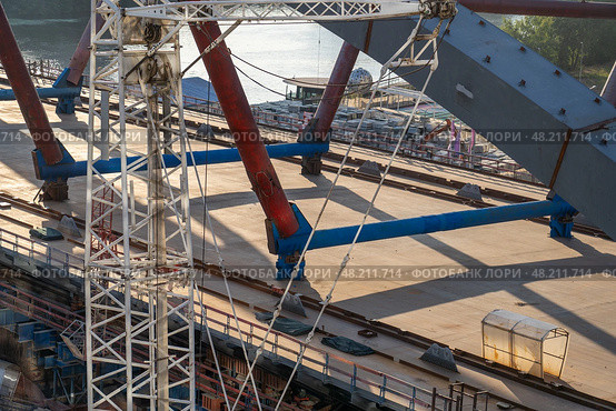 The construction of a bridge is underway with a white crane standing tall in the center of the image, supporting the steel beams overhead. Стоковое фото, фотограф Данила Большаков / Фотобанк Лори