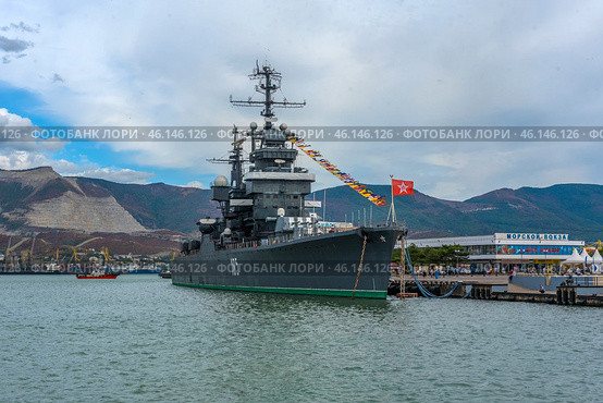 The cruiser Mikhail Kutuzov, permanently moored at the Novorossiysk embankment. This is one of the main museum ships of Russia, which is part of the Central Naval Museum.The cruiser Mikhail Kutuzov is a massive warship with the number "105" on board.It is decorated with signal flags, creating a festive atmosphere.In the background, the port of Novorossiysk, the mountains of the Markotkhsky Range and the berthing facilities are visible.The red naval flag with a white star flutters at the stern.To the right of the ship is the marine terminal, where people and city infrastructure are visible.In the distance, a small tug or boat moves along the water. (2023 год). Редакционное фото, фотограф Анна Кривицкая / Фотобанк Лори