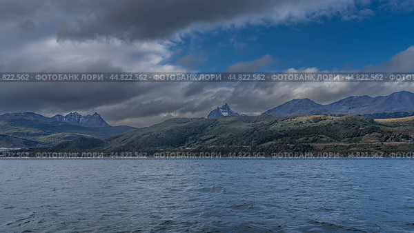 The picturesque coastline of Argentina. Snow-capped Andes mountains against the sky, clouds. At the foot of the hills are the city buildings of Ushuaia. Ripples on the ocean surface. Tierra del Fuego (2023 год). Стоковое фото, фотограф Вера Ивановна Кубарева / Фотобанк Лори