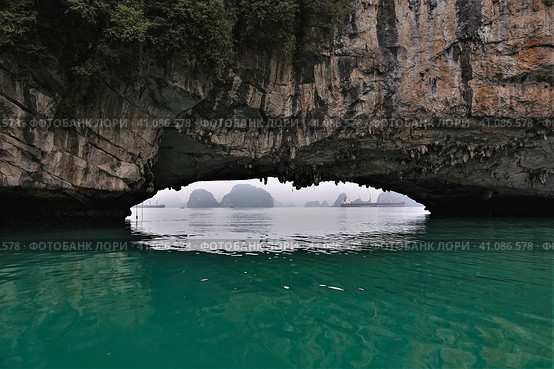 The rock hangs over the surface of the water. Through a low arched passage, you can see the islands and the ship hiding in the fog. Halong Bay. Vietnam. Close-up (2019 год). Стоковое фото, фотограф Вера Ивановна Кубарева / Фотобанк Лори