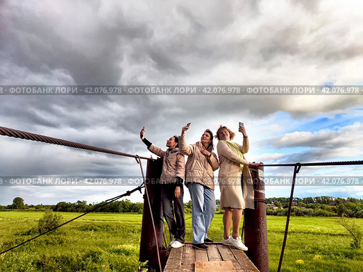 Three funny tourist girls on the old bridge take selfies against the background of an alarming dramatic sky with thunderclouds. Middle-aged women having fun outdoors. Стоковое фото, фотограф Кривошеина Елена Леонидовна / Фотобанк Лори