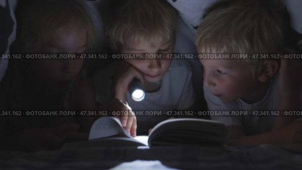 Three young children are reading a book with a flashlight under the covers at night. Стоковое видео, видеограф Mikhail Davidovich / Фотобанк Лори
