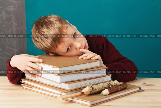 tired school boy asleep on books. little student sleeping on textbooks. Child in school uniform lies on the table with big pile of books against blue wall. School concept. Стоковое фото, фотограф Nataliia Zhekova / Фотобанк Лори