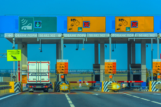 Toll booth on the M4 Don toll road in Russia. In the foreground, a truck passes through a barrier in a special lane with a green arrow, intended for transponders (automated payment system). Above each lane, there are electronic boards with payment methods (cash, card, transponder) and accessibility information. The infrastructure is clearly organized: road signs, fences, CCTV cameras, and safety signs. In the background, there is an open rural landscape with fields and trees on the horizon. (2020 год). Редакционное фото, фотограф Анна Кривицкая / Фотобанк Лори