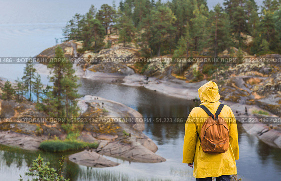 Tourism. Hiking. Ladoga skerries on Lake Ladoga. A tourist man in a yellow raincoat with a backpack on the rocky islands looks into the distance. Republic of Karelia, Russia. Стоковое фото, фотограф Photojuli / Фотобанк Лори