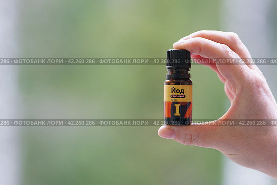 Translation of the inscription in Russian "ЙОД" - "iodine". Photography of a woman's hand holding orange vial of iodine. Window as background. Spring green color. Close up. Medical concept. Стоковое фото, фотограф YuryKara / Фотобанк Лори