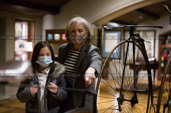 Tween schoolgirl and elderly female tutor in face masks viewing vintage bicycle in museum. Стоковое фото, фотограф Яков Филимонов / Фотобанк Лори