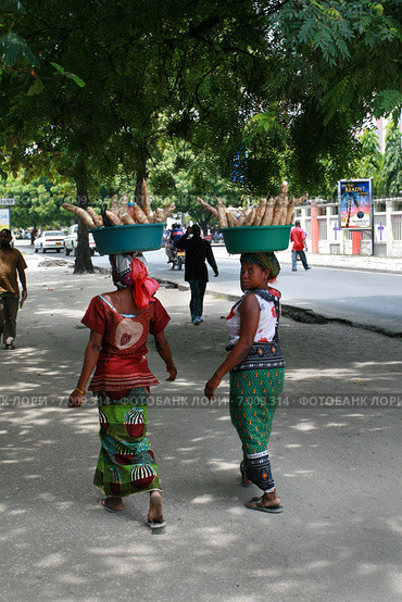 Two black women carry goods on their heads. Две танзанийки идут по дороге с грузом на голове, несут корнеплоды в зеленых, пластиковых тазах, Восточная Африка, город Дар-эс-Салам, Танзания (2008 год). Редакционное фото, фотограф Владимир Григорьев / Фотобанк Лори