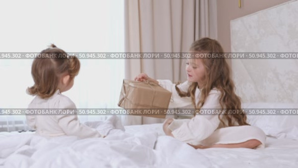Two children, little sisters sitting on a bed with white bedding, opening gift boxes wrapped in kraft paper. Holidays concept. High quality footage. Стоковое видео, видеограф Ирина Михайличенко / Фотобанк Лори
