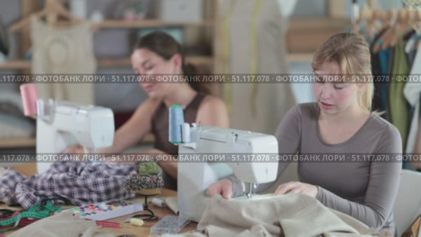 Two female tailors are sitting at a table in the workshop and sewing clothes on machines. Employees of the sewing industry make wear according to individual measurements. Стоковое видео, видеограф Яков Филимонов / Фотобанк Лори