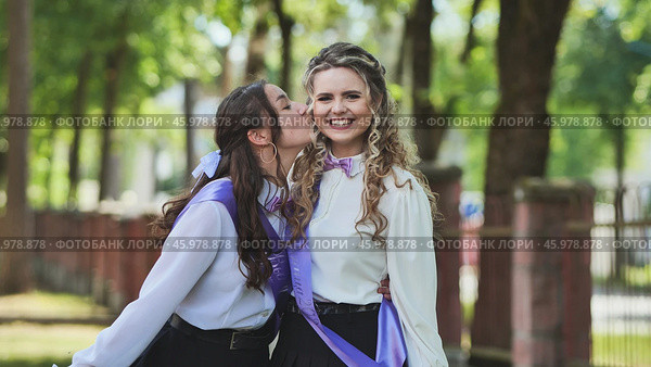 Two happy female graduates wearing sashes are kissing on the cheek, celebrating their last school day. Стоковое фото, фотограф Mikhail Davidovich / Фотобанк Лори