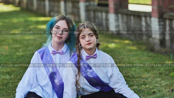 Two happy graduates celebrating their last school day, sitting on the grass in the schoolyard, wearing sashes and enjoying the moment as friends and classmates. Стоковое фото, фотограф Mikhail Davidovich / Фотобанк Лори