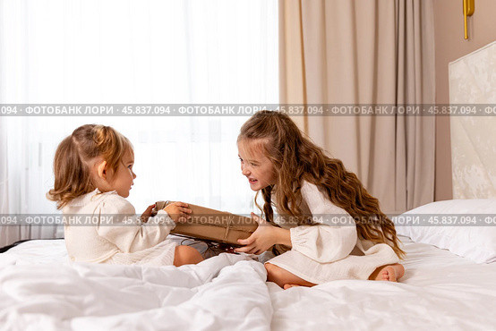 Two sisters wearing white dresses, sitting on a bed with white bedding, pulling big wrapped gift box, fighting, cannot share a present on the holiday morning at home. Стоковое фото, фотограф Ирина Михайличенко / Фотобанк Лори