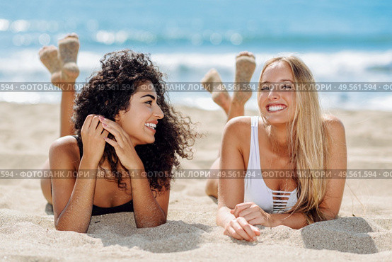 Two young women with beautiful bodies in swimwear on a tropical beach. Funny caucasian and arabic females wearing black and white swimsuits lying on the sand on the beach. Two young women with beautiful bodies in swimsuit on a tropical beach. Стоковое фото, агентство Ingram Publishing / Фотобанк Лори