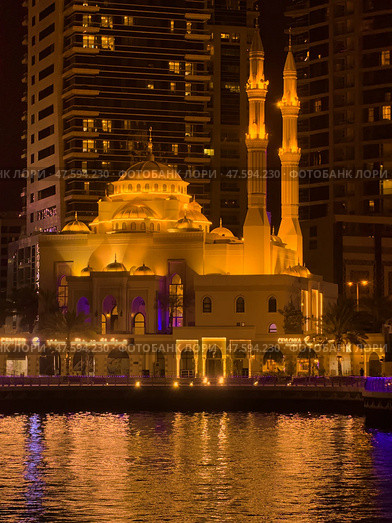 UAE, Dubai, 10 December 2024: Mosque of Muhammad Ibn Ahmad Al-Mullah against the backdrop of skyscrapers at night in Dubai Marina, yachts sailing by, reflection of night lighting on the water. Редакционное фото, фотограф Vladimir Drozdin / Фотобанк Лори