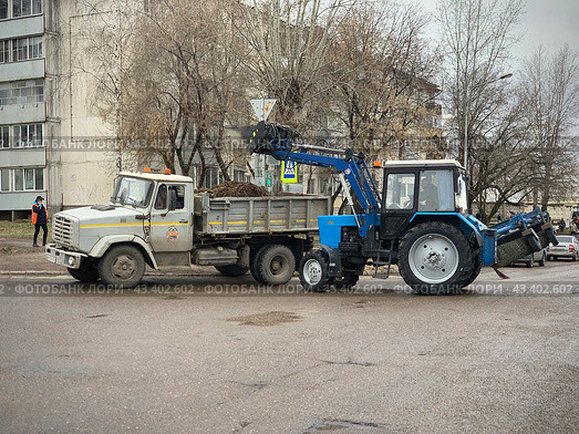 Уборочная техника трактор-экскаватор грузит мусор (прошлогоднюю листву) в грузовик на субботнике (2024 год). Редакционное фото, фотограф Ирина Тульская / Фотобанк Лори