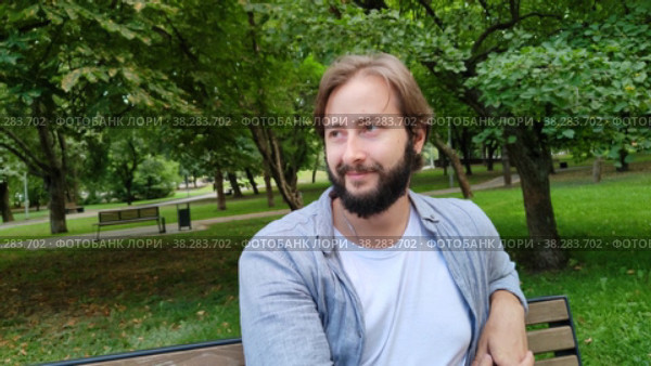 Улыбающийся красивый мужчина сидит на скамейке в городском общественном парке летним днем. Европеец отдыхает от работы и радуется жизни. Городской стиль жизни. Счастливый бородатый парень смеется. Крупный план. Smiling handsome man is sitting on bench in city public park on summer day. Caucasian man takes a break from work and enjoys life. City lifestyle. Happy bearded guy laughs. Close-up. Стоковое видео, видеограф Ирина / Фотобанк Лори