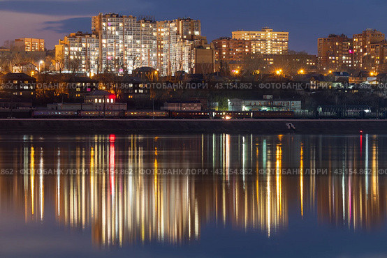 Вечерний городской пейзаж. Вид на жилые кварталы. В воде отражается свет. Вдали по берегу реки едет пассажирский поезд. Город Иркутск, Иркутская область, Россия. Река Ангара. Сибирь. Стоковое фото, фотограф Андрей Степанов / Фотобанк Лори
