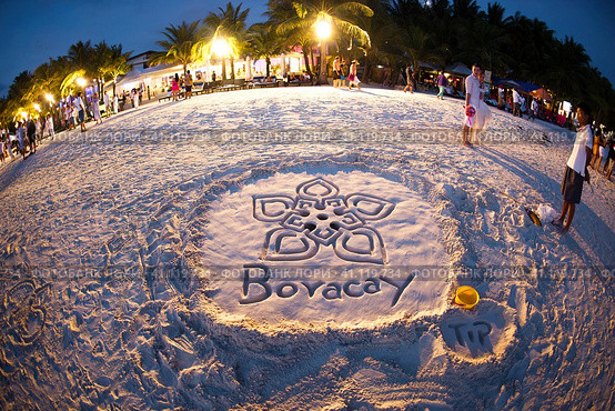 Вечерний пляж на океане. На белом песке выложена надпись Boracay и  рисунок в восточном стиле. На рисунок смотрят стоящие рядом влюбленные. Виден вечерний пляж, отдыхающие, зажженные фонари, пальмы. (2011 год). Редакционное фото, фотограф Сергей Никифоров-Гордеев / Фотобанк Лори