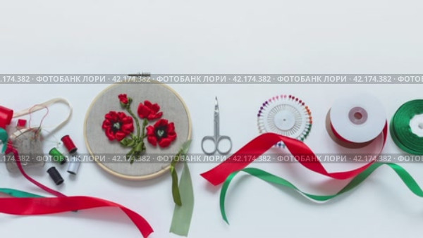 Video of handmade embroidery with satin ribbons of red spring poppies as gift. Accessories: needles, pins, threads, red and green satin ribbons on bobbins on white background. DIY concept. Flat lay, close up. Стоковое видео, видеограф Виктория Катьянова / Фотобанк Лори