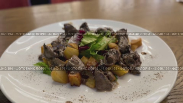 Warm salad with chicken liver in cream sauce with fried potatoes, mushrooms and mixed green salad in a white porcelain plate on a brown wooden table in an elite restaurant. Стоковое видео, видеограф worker / Фотобанк Лори