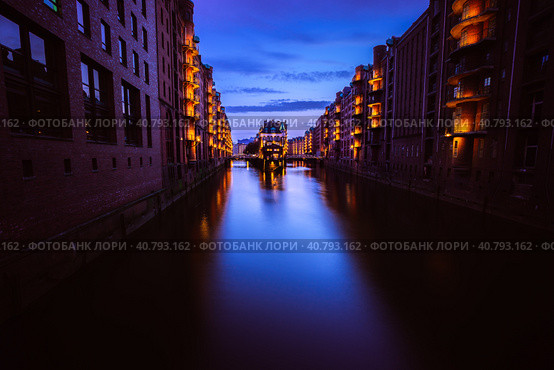 Wide perspective of Warehouse District - Speicherstadt in twilight. Tourism landmark of Hamburg. View of Wandrahmsfleet in light of lantern lamp. Port of Hamburg within the HafenCity quarter (2018 год). Стоковое фото, фотограф Igor Tichonow / Фотобанк Лори