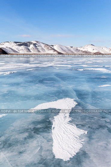Winter landscape of frozen Baikal lake on cold sunny day in February. Snow-capped hills frame iced bay, and snow crusts in form of frozen waves form from wind on blue ice surface in mouth of Anga River. Стоковое фото, фотограф Виктория Катьянова / Фотобанк Лори