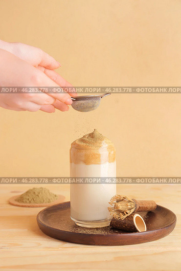 Woman adding powder onto glass with tasty dalgona hojicha latte at table. Стоковое фото, фотограф Pixel_shot / Фотобанк Лори