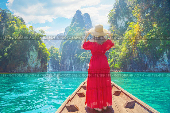Woman female traveler enjoys the breathtaking views of Cheow Lan Lake, surrounded by towering limestone cliffs and lush greenery in Khao Sok, Thailand. Wearing a vibrant red dress and wide-brimmed hat. Стоковое фото, фотограф Владимир Разгуляев / Фотобанк Лори