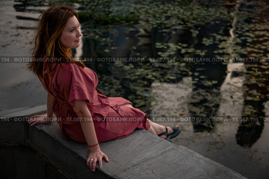 Woman in a burgundy dress sits on a stone embankment, gazing thoughtfully at the river surface with evening reflections. Стоковое фото, фотограф Софья Соломонова / Фотобанк Лори