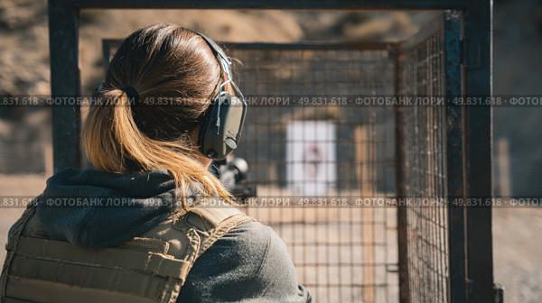Woman practicing shooting at outdoor range wearing tactical gear and ear protection, captured from behind with clear view of target downrange under natural daylight. Стоковое AI-изображение, AI-художник Катерина Бондарец / Фотобанк Лори