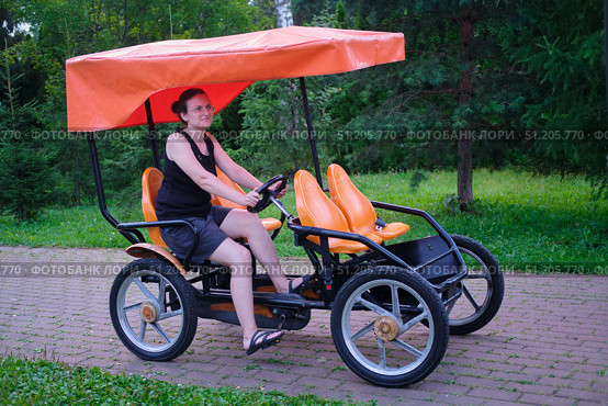 Woman riding a four-wheeled pedal bike on a paved path in a green park. Outdoor recreation and active lifestyle concept for summer. Стоковое фото, фотограф Андрей Журавлев / Фотобанк Лори