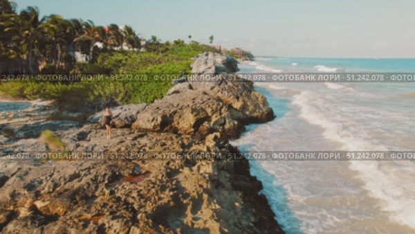 Woman walks on rocky shore path beside Caribbean Sea under morning light in Tulum, Mexico. Стоковое видео, видеограф Igor Tichonow / Фотобанк Лори
