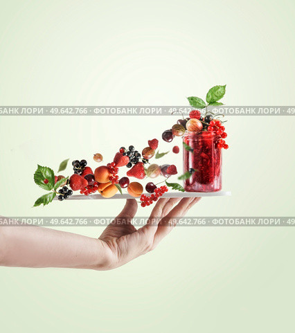 Women hand holding tray with red jam jar and various flying berries and green leaves at pale green background. Creative food levitation concept. Стоковое фото, агентство Ingram Publishing / Фотобанк Лори