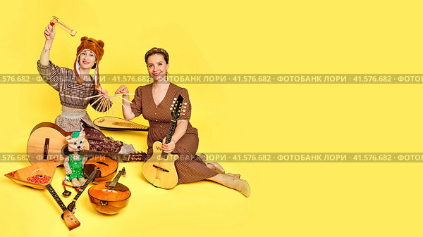 Women musicians in dresses with musical instruments on a yellow studio background. Happy artists with stringed musical instruments with smiles on faces. Стоковое фото, фотограф Андрей Журавлев / Фотобанк Лори