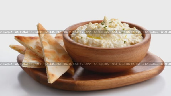 Wooden bowl brimming with creamy hummus, sprinkled with fresh herbs, resting on rustic wooden board beside golden pita triangles against crisp white surface. Стоковое AI-видео, аниматор Юрий Поздников / Фотобанк Лори