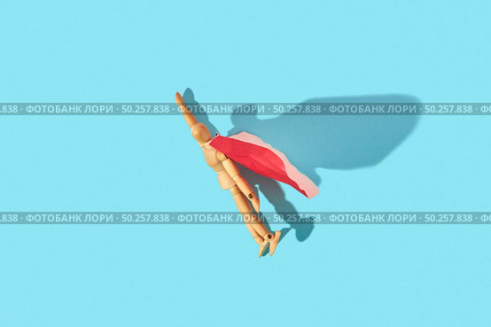 Wooden mannequin miniature doll in a red cape with raised hand flies up as a superman or superhero on a light blue background, copy space. Business success and leadeship concept.. Movable human miniature model in red cape as a superman flies up. Стоковое фото, агентство Ingram Publishing / Фотобанк Лори