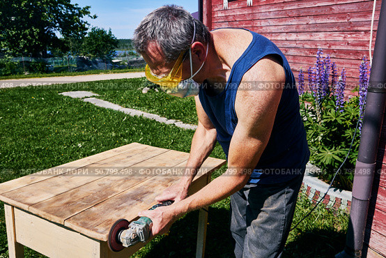 Working with handheld grinder on wooden tabletop, carpenter using protective gear during outdoor sanding in simple rural surroundings. Стоковое фото, фотограф Владимир Григорьев / Фотобанк Лори
