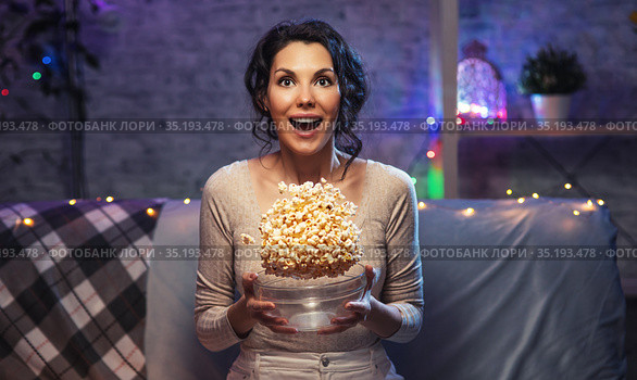 Young beautiful woman stay home, during pandemic, spends his free time watching TV on couch at home, wonder and scared, dropping popcorn. Girl watching projector movies with popcorn in evening. Стоковое фото, фотограф Сергей Тимофеев / Фотобанк Лори