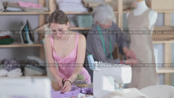 Young dressmaker cuts fabric in front of an experienced seamstress who checks a dress sample on a mannequin. Process of cutting fabric according to patterns and sewing clothes. Стоковое видео, видеограф Яков Филимонов / Фотобанк Лори