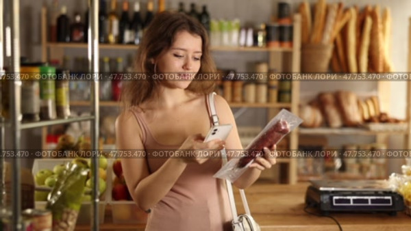Young girl using smartphone to scan package of cured sausage for detailed product information while shopping in grocery store. Concept of modern technology assisting consumers. Стоковое видео, видеограф Яков Филимонов / Фотобанк Лори