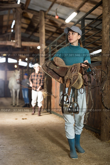Young horsewoman walking in stable with saddle in hands. Стоковое фото, фотограф Яков Филимонов / Фотобанк Лори